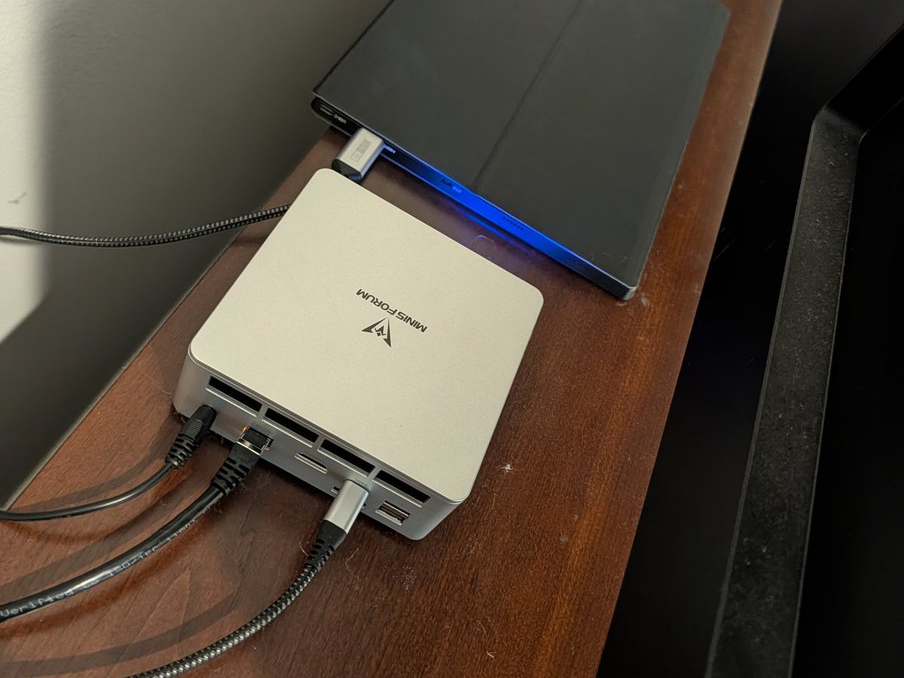 A small silver Minisforum mini PC sitting on a wooden desk, network and power cables running out of the back, a closed tablet resting behind it