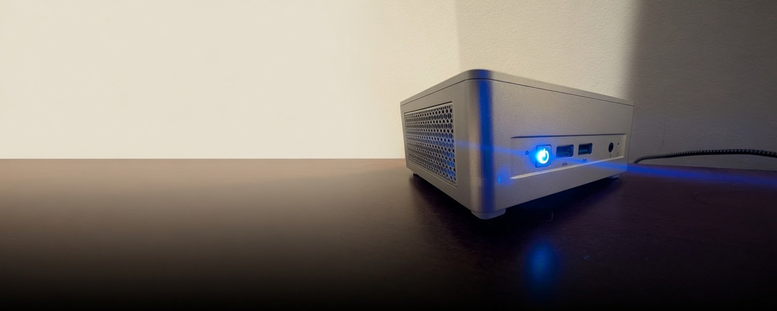 A small silver mini-PC sitting in a dark room, its front power ring lit blue, casting a faint glow across the surface it sits on